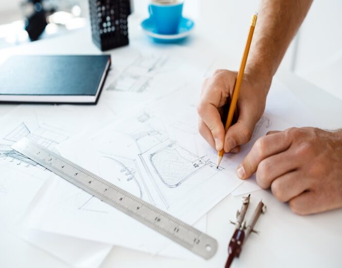 Picture of hands of young businessman holding pencil and drawing sketch at table. White modern office interior background.